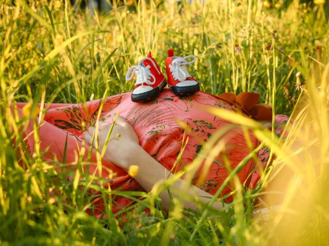 Schwangere liegt mit Kinderschuhen auf dem Bauch im hohen Gras